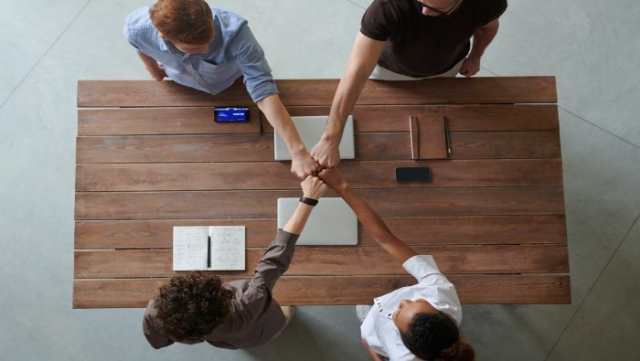 Cztery stojące nad stołem osoby dotykają się zaciśniętymi dłoni w geście współpracy